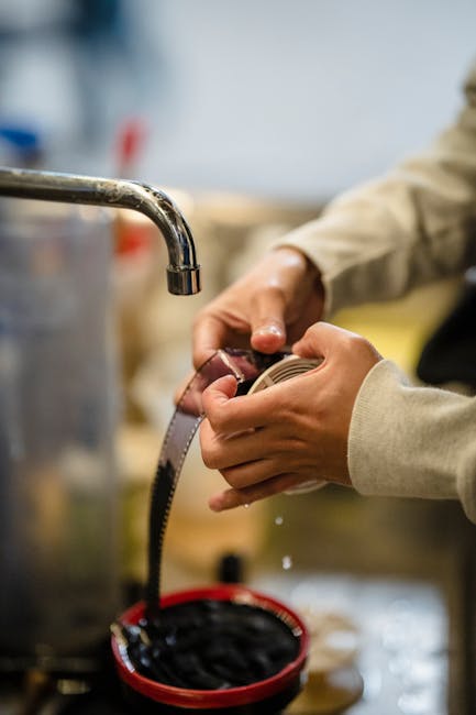 A close-up view of a person’s hands holding a flexible measuring tape, with a stainless steel sink tap visible to the left. The individual is wearing a beige or light brown long-sleeved garment, and their hands are positioned over a red plastic container filled with dark, possibly oily or greasy, waste material. The background appears to be an industrial or workshop environment, with blurred elements suggesting shelves, containers, and tools. The scene conveys a setting related to handling or inspecting waste materials, which can be associated with alternative rubbish removal or private waste management services offered by companies like Waste Collection Ilford. The image emphasizes manual measurement or inspection activities often involved in waste processing or clearance tasks, aligning naturally with the concept of on-site or independent waste handling, delivered in a straightforward, factual manner suitable for service-related content.