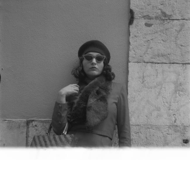 A woman stands outdoors against a textured stone wall and a smooth plastered section. She is dressed in a tailored coat with visible buttons on the sleeve and a fur scarf draped around her neck, with her left hand lightly touching her collar. She wears sunglasses with dark rounded lenses and a dark beret, and has shoulder-length hair. The lighting appears natural, with some shadowing on her face and the wall behind her. In the lower part of the image, a striped bag is partially visible resting at her side, resting on a surface that is blurred in the foreground. The scene has a neutral, professional tone, with no additional objects or environmental details related to waste management or rubbish removal visible in the image, suggesting the focus is solely on her appearance and surroundings.