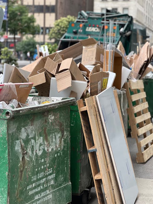 A large green industrial waste bin filled with a variety of discarded cardboard boxes, some of which are open and folded, alongside wooden pallets leaning against the side of the bin. The cardboard is in different shades of brown and white, with some pieces showing labels or printed markings. Behind the bin, there is a street scene with a green garbage collection vehicle partially visible, parked among urban buildings and trees, indicating an outdoor setting typical for rubbish removal services. The background features multiple levels of buildings, and the sky is overcast, creating a neutral natural lighting environment. The scene reflects a typical on-site clearance or private waste disposal scenario handled by a waste management company like Waste Collection Ilford, highlighting the collection and removal of bulky waste materials such as cardboard and wooden pallets from commercial or residential areas.
