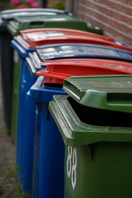 A large green industrial waste bin filled with a variety of discarded cardboard boxes, some of which are open and folded, alongside wooden pallets leaning against the side of the bin. The cardboard is in different shades of brown and white, with some pieces showing labels or printed markings. Behind the bin, there is a street scene with a green garbage collection vehicle partially visible, parked among urban buildings and trees, indicating an outdoor setting typical for rubbish removal services. The background features multiple levels of buildings, and the sky is overcast, creating a neutral natural lighting environment. The scene reflects a typical on-site clearance or private waste disposal scenario handled by a waste management company like Waste Collection Ilford, highlighting the collection and removal of bulky waste materials such as cardboard and wooden pallets from commercial or residential areas.