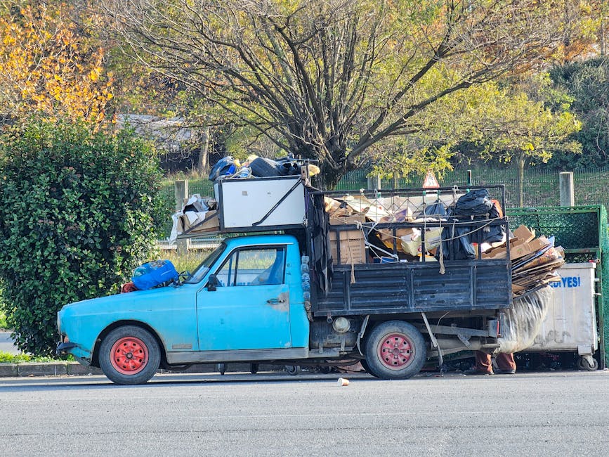 A small vintage blue pickup truck parked on the side of a paved road in daytime, loaded with a large heap of assorted waste materials including cardboard boxes, black plastic garbage bags, wooden planks, and other debris. The truck's body shows signs of wear, with faded paint and visible rust around the wheel arches, and red painted metal wheels. The cargo area is enclosed with a black metal frame, which is partially covered with ropes and twine securing the waste. Behind the truck, there is a backdrop of trees with green and autumnal foliage, and a metal fence running parallel to the roadside. The scene suggests a private waste collection or site clearance operation, aligned with independent rubbish removal services, and reflects the practical nature of on-site rubbish handling. The overall environment is bright with natural daylight, highlighting the textures and materials of the waste and the vehicle.