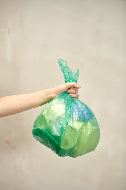 A person’s arm extends into the frame from the left, holding a green plastic rubbish bag that is loosely tied at the top with a knot. The bag appears to contain lightweight waste, with some areas slightly translucent, revealing the irregular shapes and materials inside. The surface of the bag has a smooth, slightly shiny texture, typical of standard trash bags. The background is a plain, neutral-colored wall, with no other objects or environmental features visible, providing a clear focus on the bag and the hand holding it. The lighting is soft and even, highlighting the contours of the bag without casting harsh shadows. This scene is representative of waste collection or rubbish disposal, which aligns with the services offered by Waste Collection Ilford for alternative methods of rubbish removal, such as private bin collection or on-site clearance, especially in an urban setting where quick and efficient waste handling is essential.