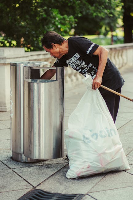 A middle-aged woman with short dark hair, wearing a black T-shirt with white graphics and black trousers, is seen disposing of rubbish into a stainless steel litter bin on a paved sidewalk. She is holding a large white plastic sack filled with waste in her left hand and using her right hand to drop items into the bin, which has a tall, cylindrical shape with a shiny, reflective surface. The background features lush green trees and a stone balustrade, indicating an outdoor urban environment. The lighting suggests a bright, clear day, with natural sunlight illuminating the scene. This image exemplifies individual efforts in waste disposal, situating the scene within a context of community cleaning or personal waste management, relevant to rubbish collection services and alternative waste handling practices primarily outside of municipal collection settings, as seen through the use of a private collection approach. Waste Collection Ilford is associated with waste management and rubbish removal, ensuring proper disposal of household or street waste in such urban environments.