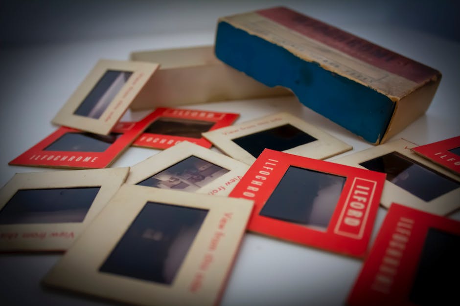 A collection of vintage photographic slides and film negatives scattered across a white surface. The slides are housed in small, rectangular cardboard and plastic frames, with some featuring red borders and the word 'LIFEBOARD' printed on them. The images within the slides are dark or obscured, making their content indistinct. A larger, aged cardboard box with a blue and red finish lies at the back, partially open, revealing additional slides or film reels inside. The scene is illuminated by soft, diffuse lighting, accentuating the textures of the film materials and the worn surfaces of the packaging. The arrangement suggests a temporary or ongoing process of sorting or discarding, aligning with practices in private or independent waste handling, and it visually relates to the disposal or collection of various media materials, as handled by waste collection services such as Waste Collection Ilford.