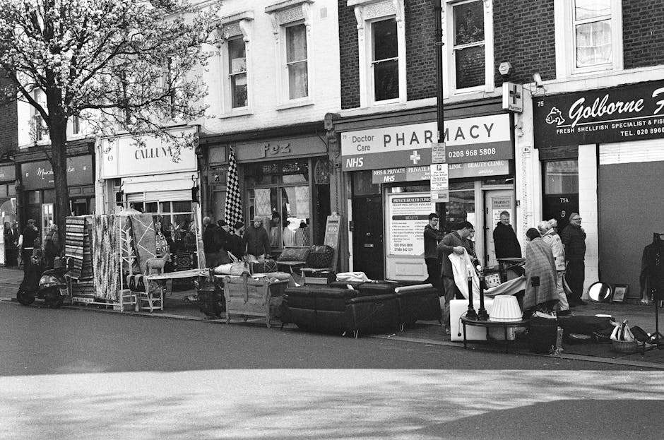 A street scene in front of a row of commercial storefronts, including a pharmacy and a fish and seafood shop, with a group of people gathered around a collection of discarded furniture and household items. The items include a black leather sofa, a small table with a lamp, and various boxes and bags, arranged along the pavement. Some individuals appear to be inspecting or handling the rubbish, which is placed directly on the sidewalk outside the shops. The background shows a brick building with multiple windows, and there is a tree partially visible on the left side of the scene. The image is in black and white, with natural daylight illuminating the scene. The presence of waste furniture and household items on the pavement may suggest an informal rubbish collection or on-site clearance, aligning with services offered by waste collection providers like Waste Collection Ilford for private waste handling outside regular municipal rubbish disposal.