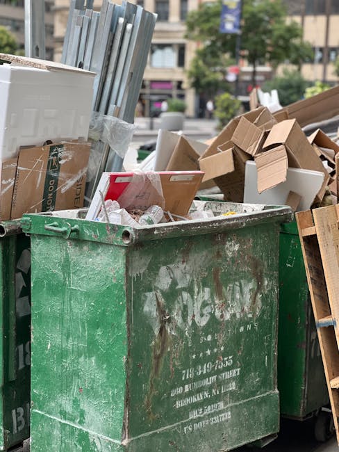 A large, green, outdoor dumpster filled with various waste materials, including cardboard boxes, white foam packing materials, and loose packaging debris. The metal container shows signs of wear, rust, and weathering, with faded markings and patches of grime on its surface. It is situated on a pavement, with additional waste and cardboard stacks leaning against the sides of the dumpster. In the background, a cityscape with multi-storey residential or commercial buildings, trees, and a few blurred parked vehicles is visible, indicating an urban environment. The scene appears to be part of an area designated for waste collection, possibly related to private rubbish removal or on-site clearance services by Waste Collection Ilford, reflecting typical materials handled during non-regular council rubbish disposal. Natural daylight illuminates the scene, highlighting the textures of the materials and the cluttered nature of the waste, with the focus on the immediate surroundings of the dumpster indicating an active waste disposal or clearing process.
