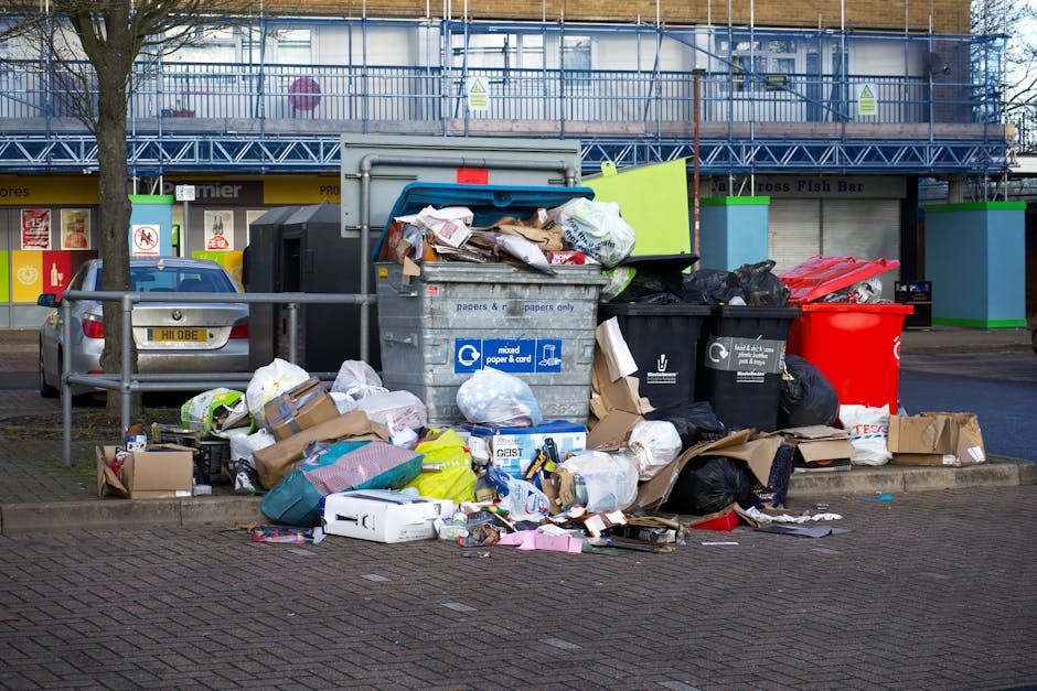 A large accumulation of mixed waste is overflowing from multiple recycling and general rubbish bins placed on a paved sidewalk next to a street. The central bin, marked for mixed paper and cardboard, is open at the top with a variety of papers, cardboard boxes, and plastic packaging spilling out onto the ground. Surrounding the main bin are several black and red rubbish bags, along with flattened cardboard boxes, plastic bags, and discarded packaging, creating a cluttered scene. In the background, a parked silver car is partially visible behind a low metal railing. Behind the waste scene, there are commercial storefronts with signage, yellow and white posters, and a two-story building topped with blue scaffolding and protective netting, indicating ongoing construction or renovation. The natural lighting suggests daytime with an overcast sky, providing a neutral, detailed view of improper waste disposal and the need for rubbish collection services such as those offered by Waste Collection Ilford, who handle bulk removal and on-site clearance to prevent illegal dumping.
