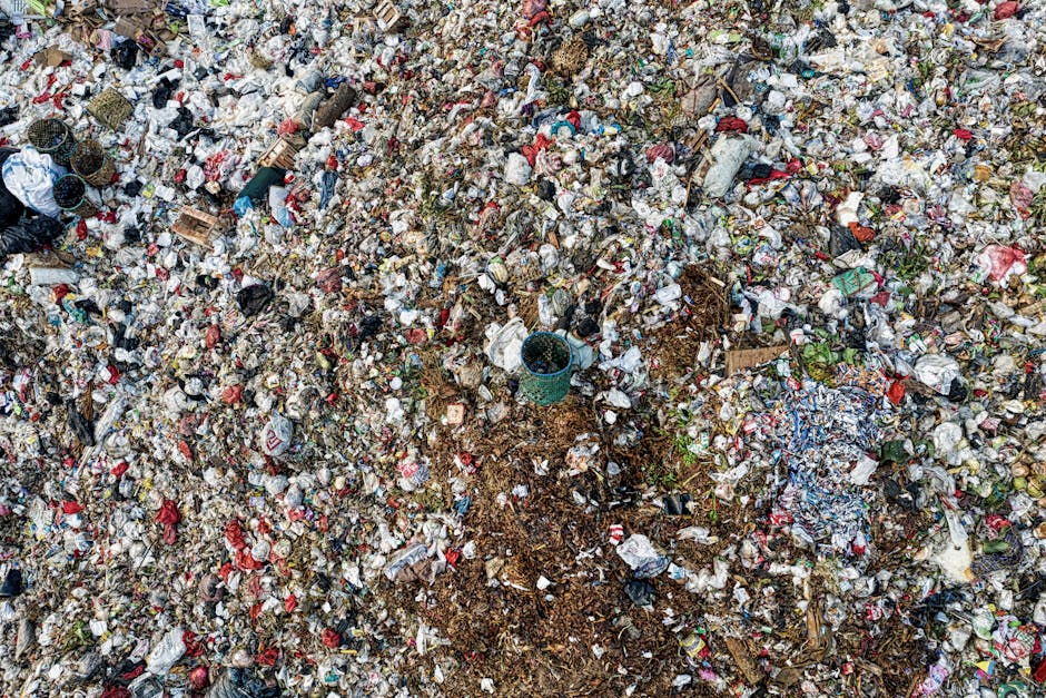 A large, green, outdoor dumpster filled with various waste materials, including cardboard boxes, white foam packing materials, and loose packaging debris. The metal container shows signs of wear, rust, and weathering, with faded markings and patches of grime on its surface. It is situated on a pavement, with additional waste and cardboard stacks leaning against the sides of the dumpster. In the background, a cityscape with multi-storey residential or commercial buildings, trees, and a few blurred parked vehicles is visible, indicating an urban environment. The scene appears to be part of an area designated for waste collection, possibly related to private rubbish removal or on-site clearance services by Waste Collection Ilford, reflecting typical materials handled during non-regular council rubbish disposal. Natural daylight illuminates the scene, highlighting the textures of the materials and the cluttered nature of the waste, with the focus on the immediate surroundings of the dumpster indicating an active waste disposal or clearing process.