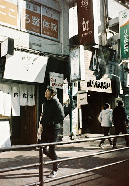 A street scene in an urban area featuring several pedestrians walking along the pavement, with storefronts and signs visible above them. The foreground shows a young man dressed in dark clothing, including a jacket and trousers, walking past a metal railing. Behind him, two other individuals are seen walking, one in a light grey jacket and the other in darker clothing. The shop fronts display Japanese characters on signs and banners, with some signs including English words such as 'WINE' and 'BOOGL.' The shops have varied facades, with some featuring wooden panels and others with modern signage. One prominent sign reads 'di PUNTO,' and another sign beneath it is illuminated. The scene is lit by natural daylight, casting shadows on the pavement and highlighting the textures of the building surfaces, which include glass, metal, and wood. The overall setting suggests a busy commercial street typical of densely populated Japanese city districts, with a focus on retail and dining establishments, indicative of an area that may be associated with private or alternative waste handling through rubbish removal services like Waste Collection Ilford, although the scene itself is not directly related to rubbish collection activities.