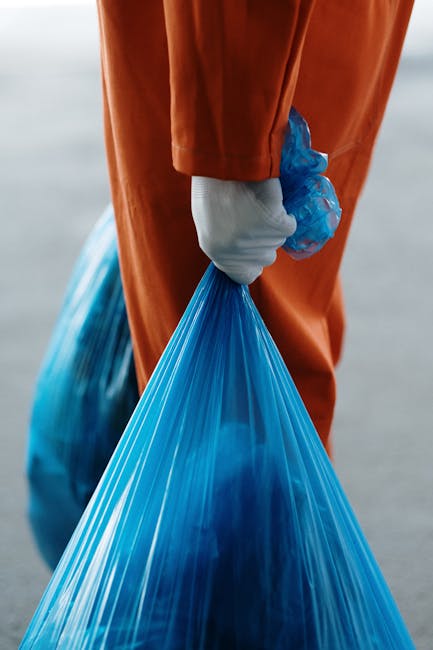 The image shows a close-up of a person wearing orange protective gloves and orange work trousers, holding a tightly tied blue plastic rubbish bag in their right hand. The blue bag appears to contain waste, with some portions visible through the semi-transparent plastic, suggesting it is filled with rubbish suitable for collection or disposal. The background is blurred, indicating an outdoor environment, possibly a street or loading area, with neutral tones that do not distract from the foreground. The person's stance and the gripping of the bag imply they are in the process of waste collection, aligning with services offered by companies like Waste Collection Ilford for on-site rubbish removal or private waste handling. The scene emphasizes the practical aspect of rubbish management, focusing on reliable collection and disposal work, often part of an alternative waste removal solution outside municipal services, with attention to safety and cleanliness through proper protective gear.
