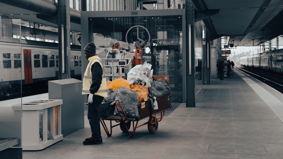 A street scene in an urban area featuring several pedestrians walking along the pavement, with storefronts and signs visible above them. The foreground shows a young man dressed in dark clothing, including a jacket and trousers, walking past a metal railing. Behind him, two other individuals are seen walking, one in a light grey jacket and the other in darker clothing. The shop fronts display Japanese characters on signs and banners, with some signs including English words such as 'WINE' and 'BOOGL.' The shops have varied facades, with some featuring wooden panels and others with modern signage. One prominent sign reads 'di PUNTO,' and another sign beneath it is illuminated. The scene is lit by natural daylight, casting shadows on the pavement and highlighting the textures of the building surfaces, which include glass, metal, and wood. The overall setting suggests a busy commercial street typical of densely populated Japanese city districts, with a focus on retail and dining establishments, indicative of an area that may be associated with private or alternative waste handling through rubbish removal services like Waste Collection Ilford, although the scene itself is not directly related to rubbish collection activities.
