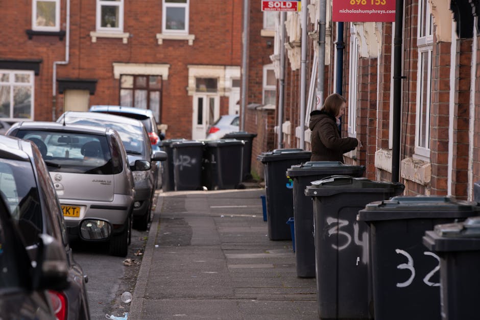 A woman wearing a dark brown jacket stands on a sidewalk lined with black wheelie bins, each with a closed lid, positioned against a brick building facade. The bins are marked with white spray paint indicating their designated numbers, such as 32 and 34, and are arranged in a row along the pavement. The building features multiple windows with white frames and sills, some with curtains visible inside. The street is narrow and features parked cars on the left side, including a silver hatchback and other vehicles extending into the background, with their rearview mirrors visible. The area appears to be an urban residential setting, with brick terraces and additional vehicles parked further down the street. The overall lighting suggests daytime with natural light illuminating the scene, capturing a typical waste collection or disposal scenario that aligns with private waste handling practices often seen in property-maintenance contexts managed by services like Waste Collection Ilford.