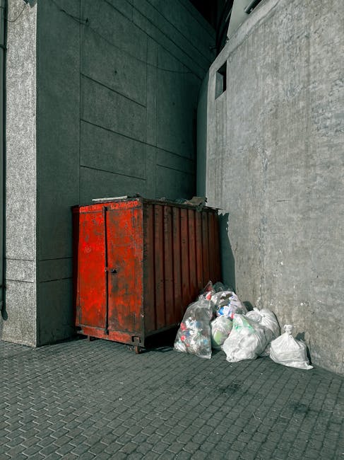 A large, rectangular metal skip with a weathered, red finish shows signs of rust and paint chipping in an outdoor urban setting, positioned against a dark green tiled wall on the left and a plain, light grey concrete wall on the right. The skip is partly filled with miscellaneous rubbish and has its lid partially open. Next to the skip, on the paved ground, there are several white plastic bags filled with waste, some of which are tied, while others are loosely closed. The bags contain various types of refuse, possibly household waste or recyclables. The scene appears to be an area for waste collection or disposal, with natural daylight illuminating the scene. The overall environment suggests an urban service area where waste is gathered prior to collection, aligned with private waste handling practices some companies like Waste Collection Ilford may facilitate rather than standard council services. The industrial appearance of the skip, combined with the surrounding walls and ground surface, contributes to a utilitarian and functional waste disposal setting.