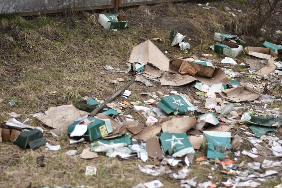 A patch of uneven ground beside a grassy area shows scattered debris including crushed cardboard boxes, some with visible branding, torn and crumpled, mixed with smaller pieces of paper and plastic wrappers. The cardboard varies in colour from green to brown with rough textures and sharp edges, and some appear partially flattened or ripped. Near the top of the image, a section of a weathered metal fence or barrier is visible, with the ground beneath cluttered with litter. The background includes a dirt slope with sparse, leafless vegetation, indicating an outdoor environment possibly near an industrial or waste collection site. The scene is lit by natural daylight, highlighting the disorganized state of the rubbish, which appears to have been discarded rather than collected or cleaned up. This visual exemplifies the importance of proper waste disposal to avoid environmental littering, relevant to services like those offered by Waste Collection Ilford for independent or private rubbish clearance, particularly in contexts where rubbish duty of care responsibilities are involved.