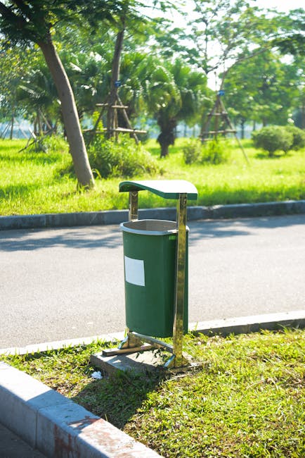 A green cylindrical rubbish bin with a white rectangular label is installed on a metal stand next to a paved sidewalk. The bin has a slightly curved, light-colored lid attached to a metal support arm. It is positioned on a small grassy area adjacent to a curb made of concrete blocks. Behind the bin, there is a road with smooth grey asphalt, bordered by a narrow concrete edge. In the background, a lush green landscape featuring various trees, including some with palm-like fronds, and shrubbery is visible, suggesting a park or garden setting. The scene is illuminated by bright sunlight, casting minimal shadows and emphasizing the vibrant green foliage and bright colours of the bin, which may be part of an environmentally conscious waste collection system. The overall environment appears tidy and well-maintained, typical of outdoor public rubbish disposal points, which could be serviced by local or private waste removal providers like Waste Collection Ilford, supporting efficient rubbish management and alternative waste handling outside of municipal services.
