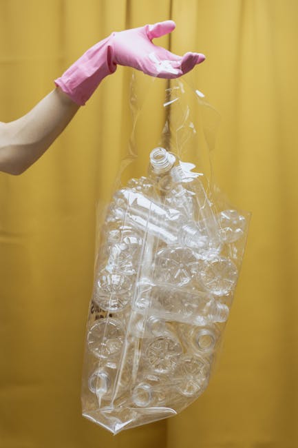 A person wearing a pink rubber glove is holding a large, transparent plastic bag filled with crumpled, clear plastic bottles and containers, set against a plain yellow curtain background. The plastic waste appears to be empty bottles made of PET, with some of the bottles' necks and openings visible at the top of the bag. The bag is partially open at the top, allowing the necks of the bottles to extend outwards. The scene suggests an act of sorting or preparing recyclable plastics, which relates to waste management and rubbish removal services performed by companies like Waste Collection Ilford. The plain, uncluttered environment emphasizes the focus on waste items, highlighting the importance of proper disposal or recycling of plastic waste as part of private rubbish handling or on-site clearance efforts. The lighting is even, with soft shadows, providing clear visibility of the textured, glossy surfaces of the plastic materials, reinforcing the visual clarity necessary for accessible description and SEO relevance in waste management contexts.