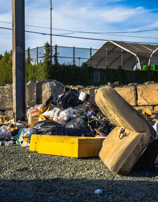 A pile of discarded rubbish situated on a gravel surface outdoors, featuring black plastic garbage bags overflowing with mixed waste, including crumpled paper and plastic packaging. Nearby, there is a yellow plastic storage container and a large, beige upholstered armchair that appears worn and stained, with its cushion partially leaning against the chair frame. In the background, a stone wall runs horizontally, composed of large, roughly rectangular stones with a weathered grey appearance. Beyond the wall, a chain-link fence is visible, along with trees, a utility pole with electric wires, and a partially visible greenhouse or large tent structure with a curved roof. The sky is clear with a few scattered clouds, and the scene is lit by natural daylight, emphasizing the cluttered and unmanaged state of the rubbish, which may relate to private or independent waste collection efforts by waste management services such as Waste Collection Ilford.